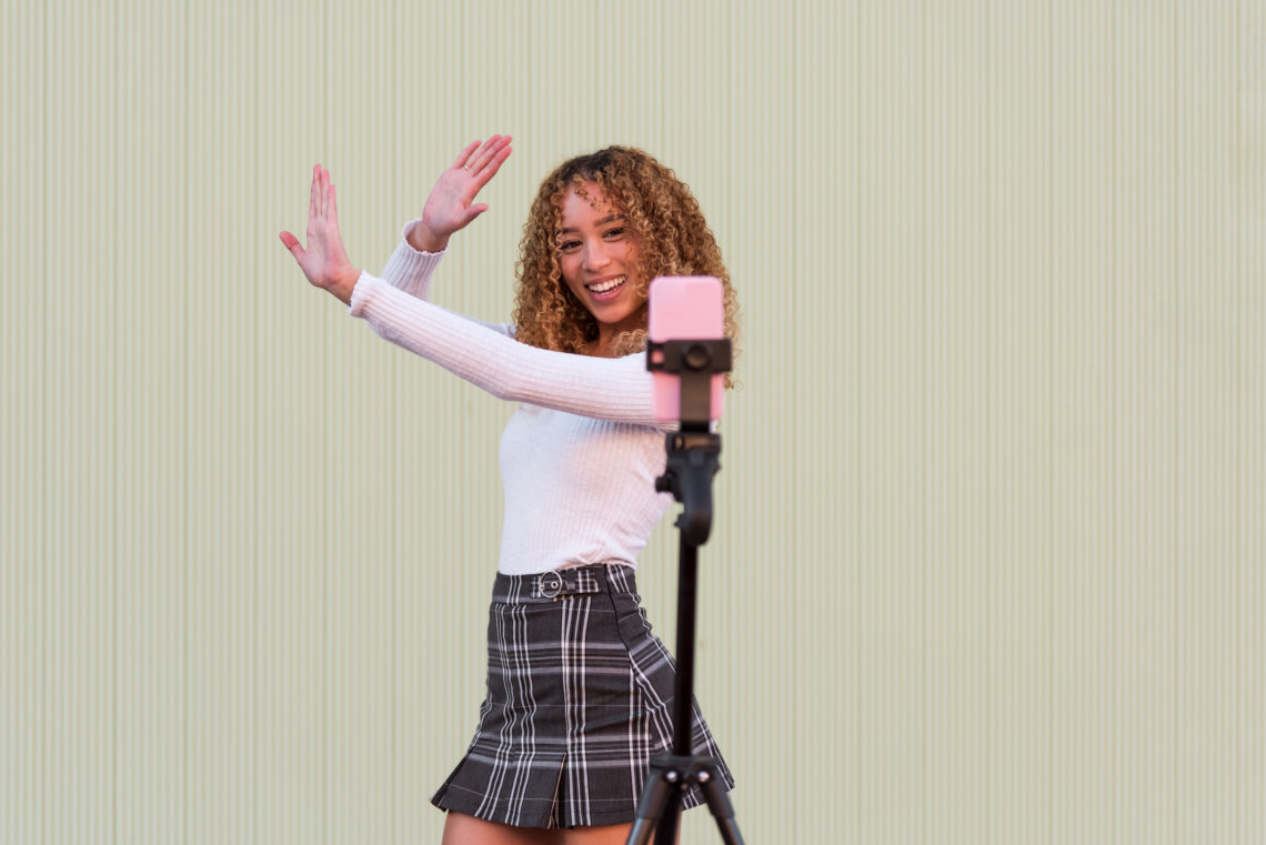 Positive Hispanic female blogger in stylish wear with curly hair dancing and recording video for Tik Tok on cellphone while standing near wall on s...