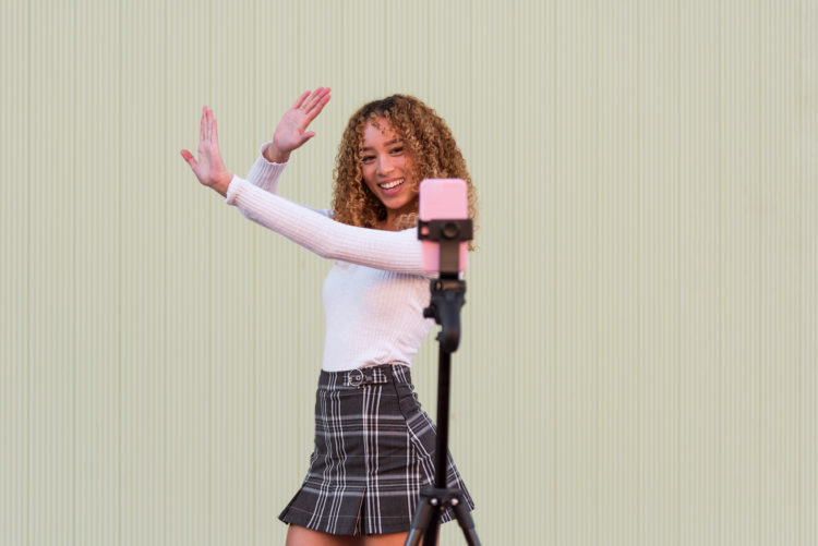 Positive Hispanic female blogger in stylish wear with curly hair dancing and recording video for Tik Tok on cellphone while standing near wall on s...