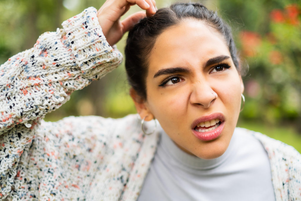 Young woman with hair pulled back and wearing speckled cardigan scratching her head and looking confused