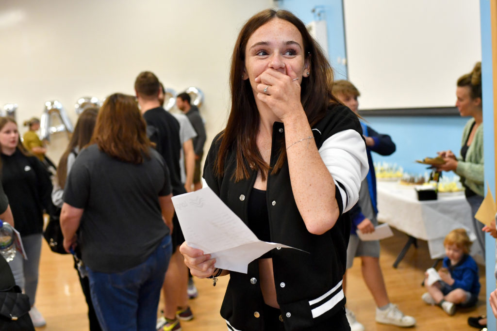 Female student in black and white baseball jacket hold GCSE results and hold hand to mouth with happy expression looking off camera. She stands in a hall with other students nehind her.