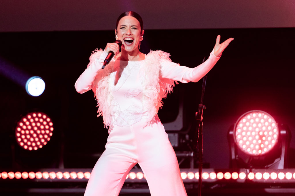 Blanca Paloma in white feathered top and white trousers holding microphone with other arm outstretched performing on stage