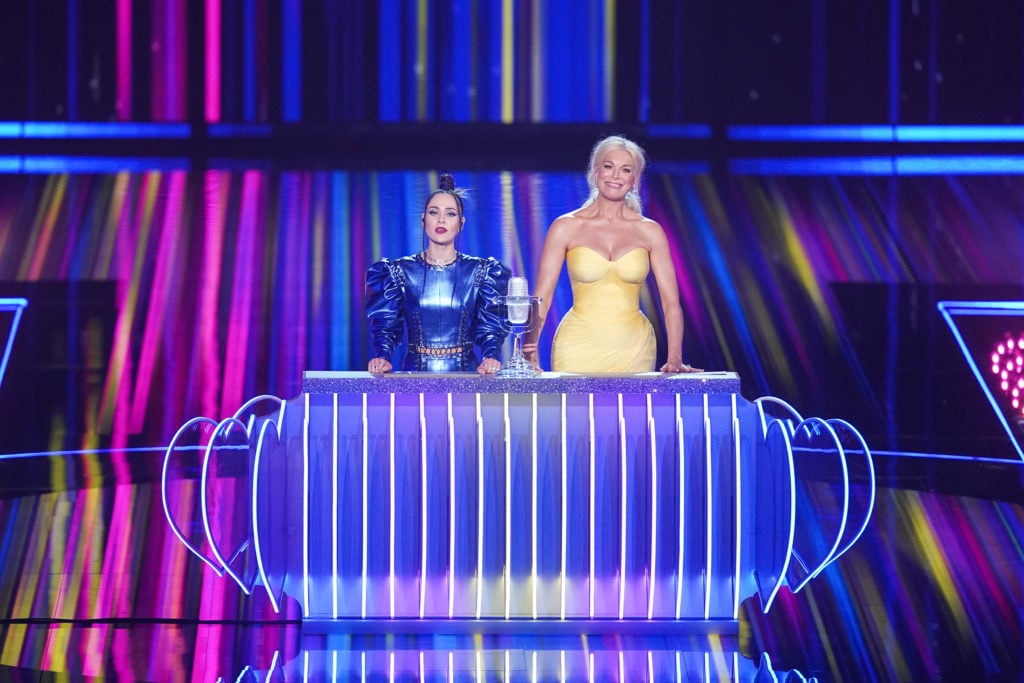 (l-r) Julia Sanina in blue metallic dress next to Hannah Waddingham in yellow dress hosting on Eurovision stage, stoof behind a desk.