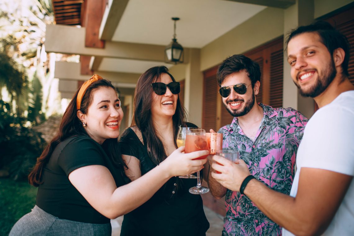 group of four friends made up of two young women and men stood together outside clinking various drinks, smiling and laughing