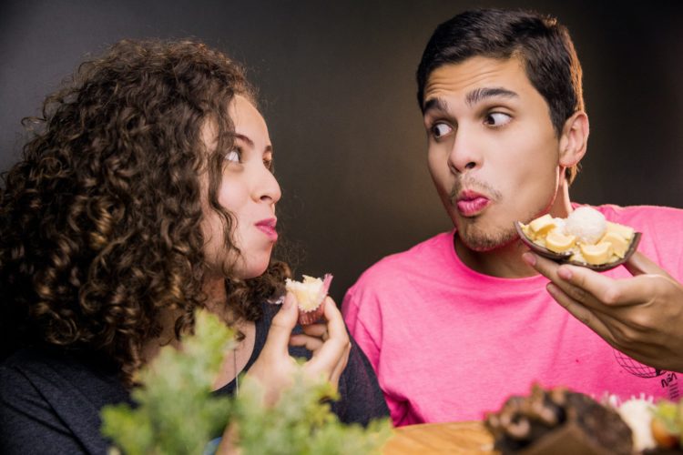 A young girl and man looking at each other whilst eating food. They hold some food in their hand whilst chewing.
