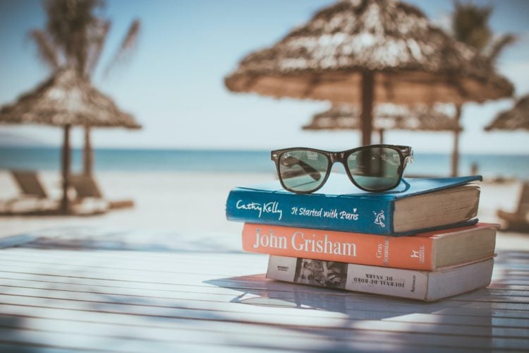 sunglasses balances on three books on a wooden table on sunny beach.