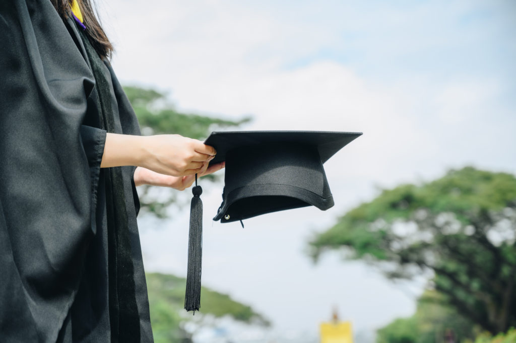 young woman in black academic dress holding cap out in front of her on sunny day