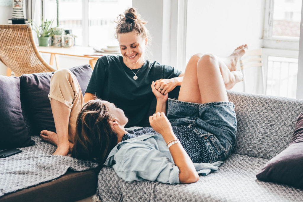 Two women lying on a grey sofa. One is in black tshirt with camel trousers, the other is lying upside down in blue denim dungarees with blue shirt