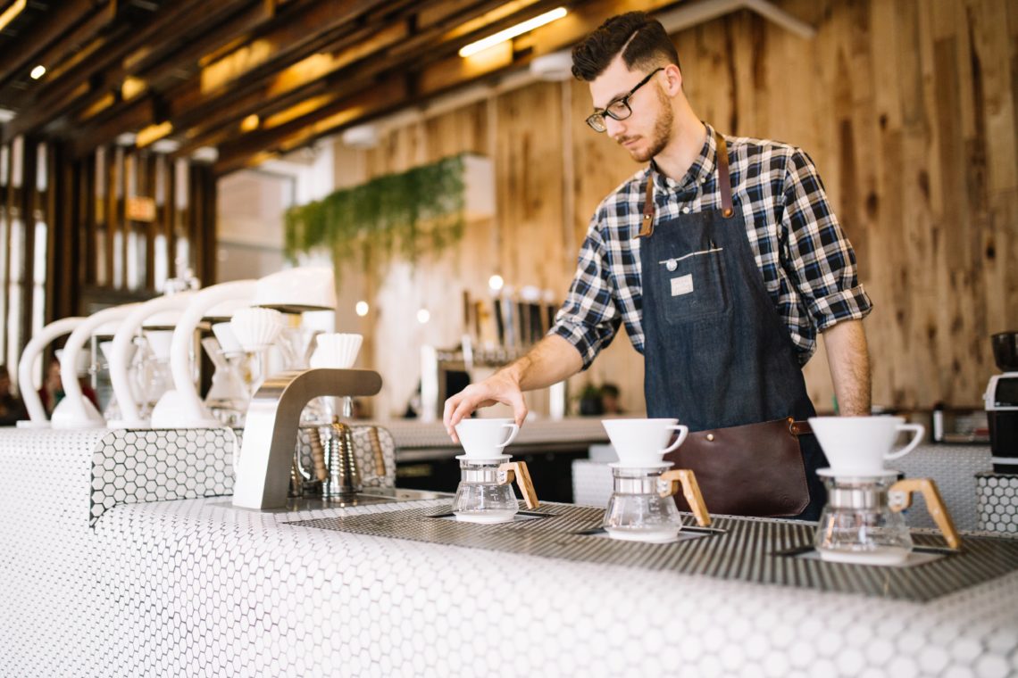 man in blue and white plaid button up shirt and apron standing in front of coffee shop counter with cups and machines