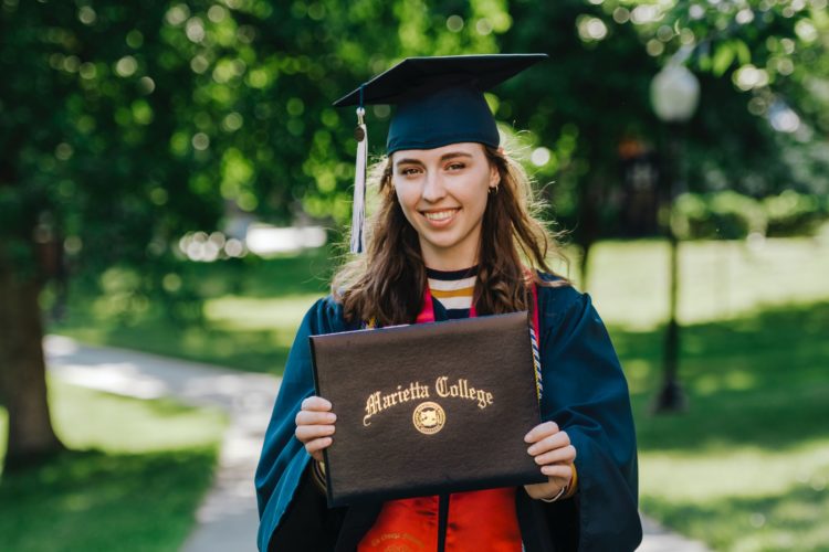 woman in academic dress wearing academic hat holding up degree whilst stood outside