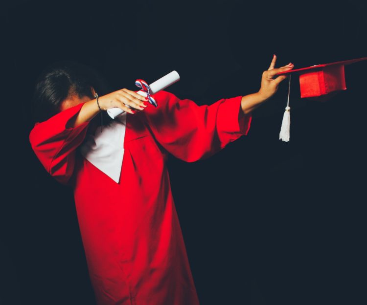 person wearing red graduation dress dabbing holding scroll in one hand and cap in another against a black background