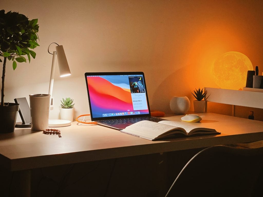 macbook pro on white wooden desk with desk lamp on and book open