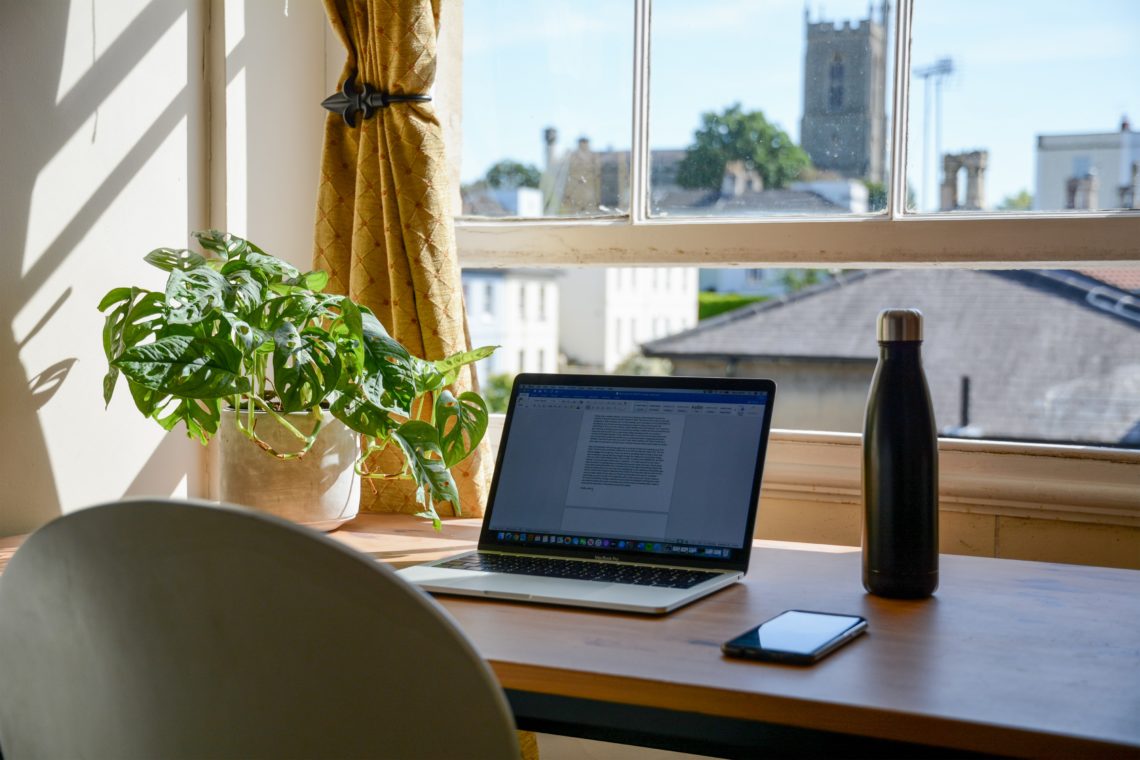 ope laptop on wooden desk next to water bottle in front of window on sunny day.
