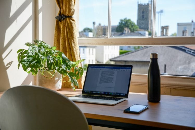 ope laptop on wooden desk next to water bottle in front of window on sunny day.