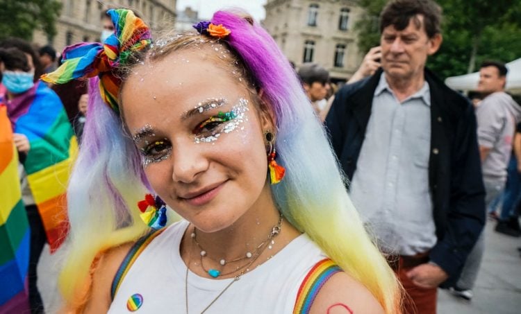 woman in white tank top with purple and yellow hair tie wearing various rainbow accessories with people behind her