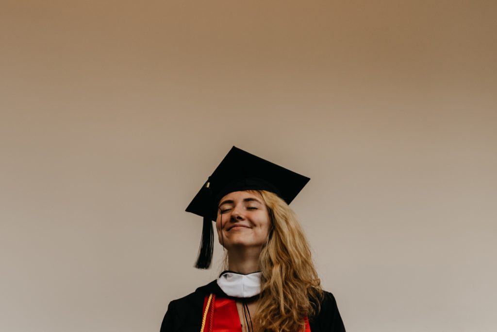 young woman in black and red graduate dress stood against white wall